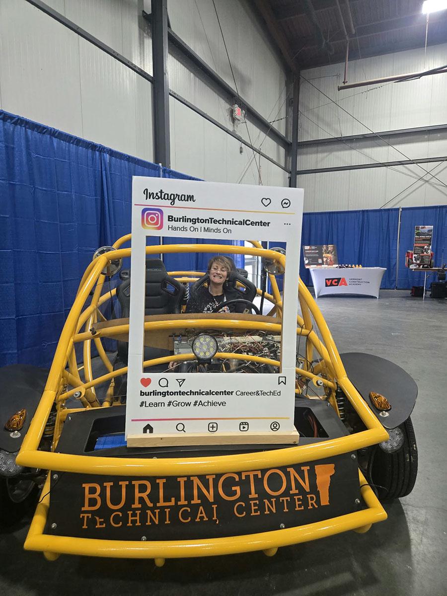 A person seated in a yellow Burlington Technical Center vehicle behind an Instagram-shaped display frame